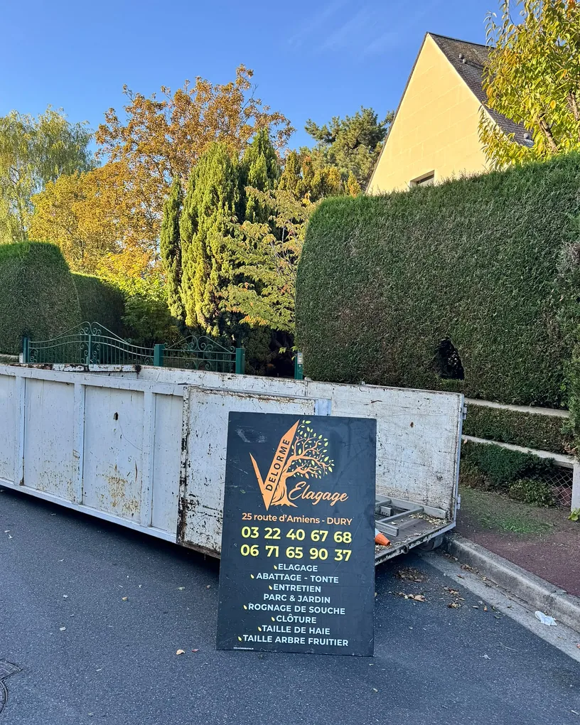 Panneau Delorme Élagage listant élagage, abattage, tonte, entretien parc et jardin, taille de haie, devant une benne de chantier
